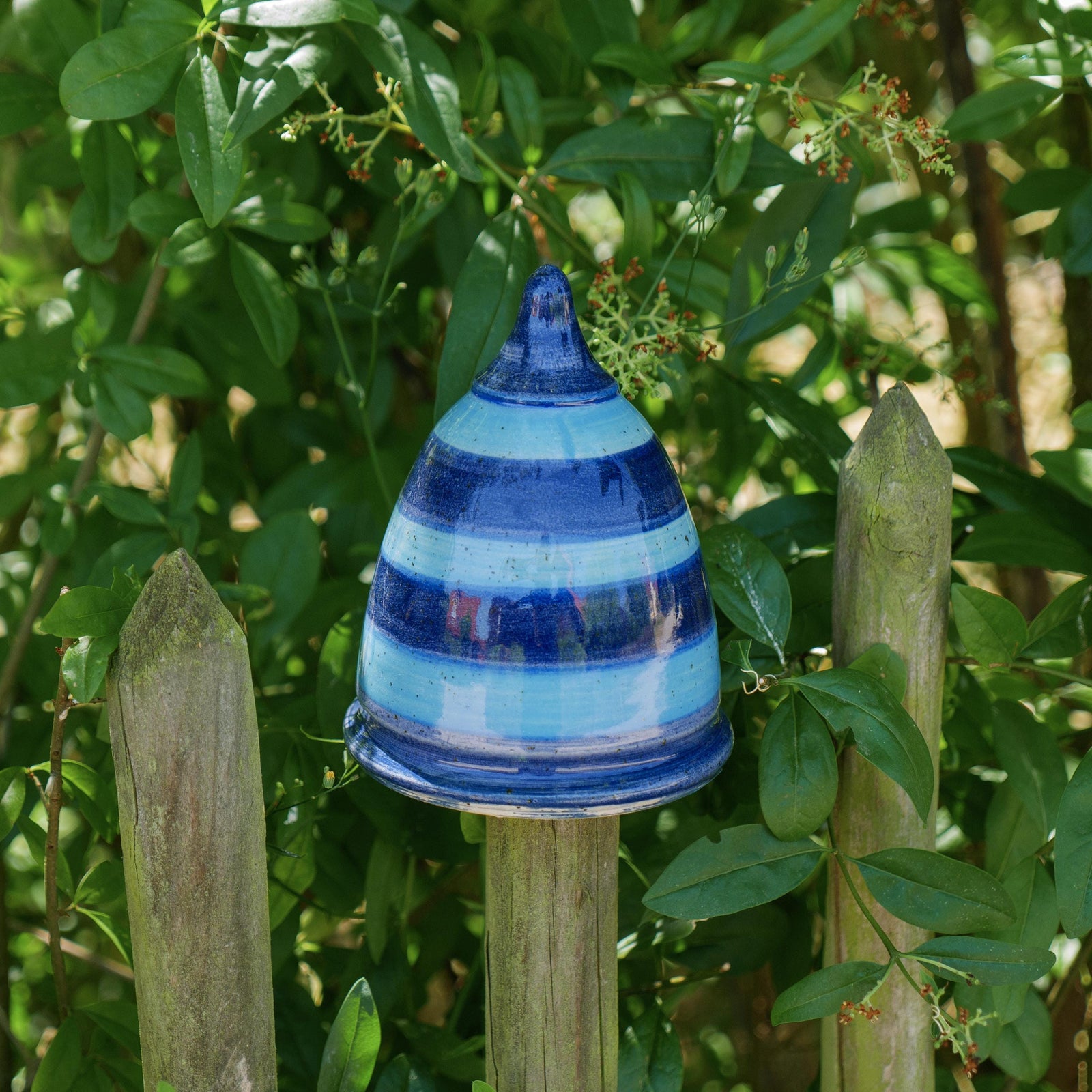 Handgefertigter blau-gestreifter Keramik-Pfostenhocker im Garten, glasiert in mehreren Blautönen mit horizontalen Streifen. Kegelförmige Gartenspitze, ca. 12 cm hoch, mit einer 5–6 cm großen Öffnung unten – passend für Zaunpfähle oder Holzstäbe. Das Stück steht auf einem Gartenzaun zwischen grünen Pflanzen im Freien.
