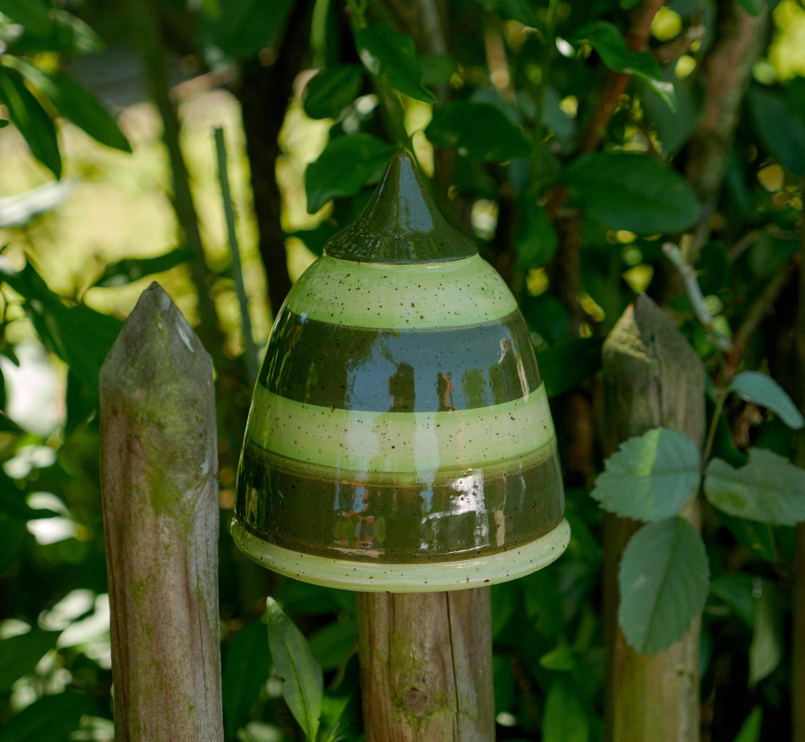 Handgefertigtes Keramik-Gartenornament in abgerundeter Kegelform mit glänzender grün-brauner Streifenglasur, auf einem Holzzaun zwischen Blättern befestigt.