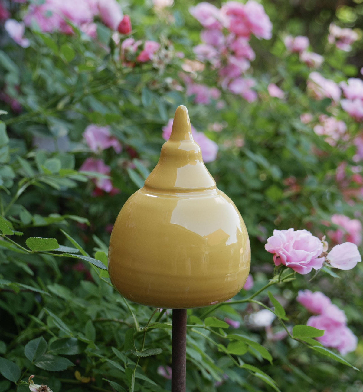 Handgefertigte Keramik-Gartenstele in glänzendem Senfgelb mit abgerundeter Kegelform, aufgestellt zwischen blühenden rosa Rosen