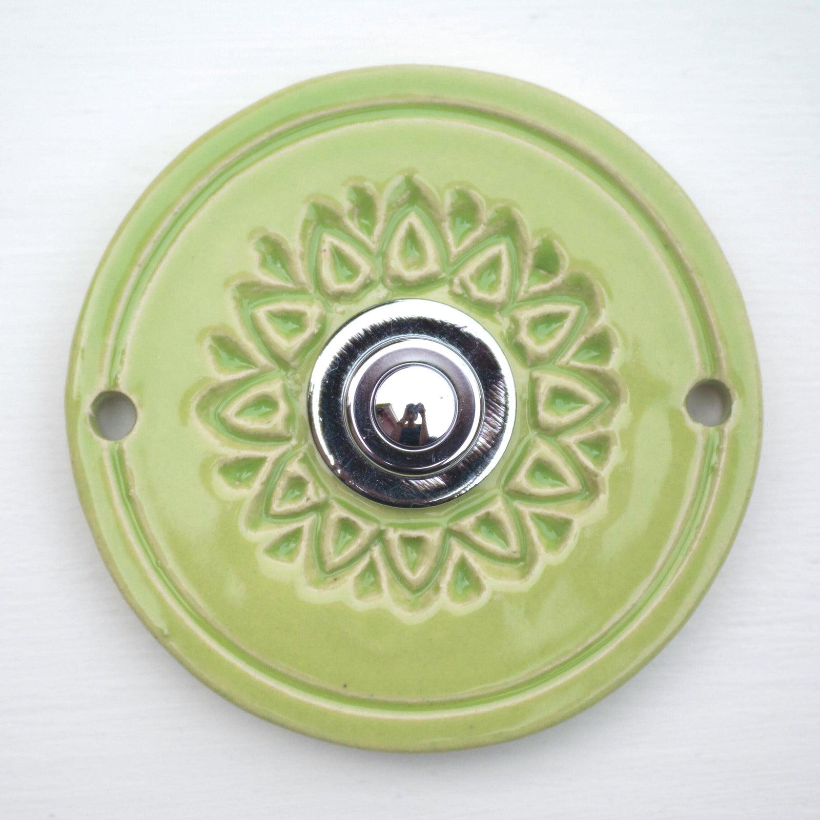 Runde dekorative Klingel mit geprägtem Blumen-Sternmuster auf hellgrüner Platte und silbernem Druckknopf in der Mitte.