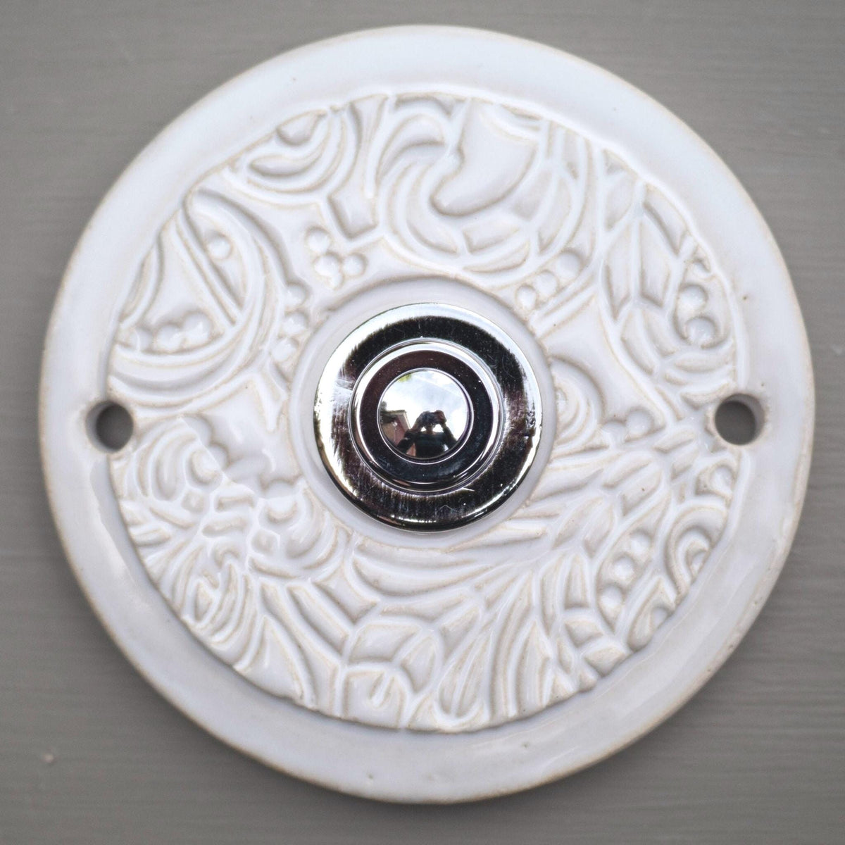 Runde dekorative Klingel mit reinweißem Ornamentmuster auf grauer Platte und goldenem Druckknopf in der Mitte