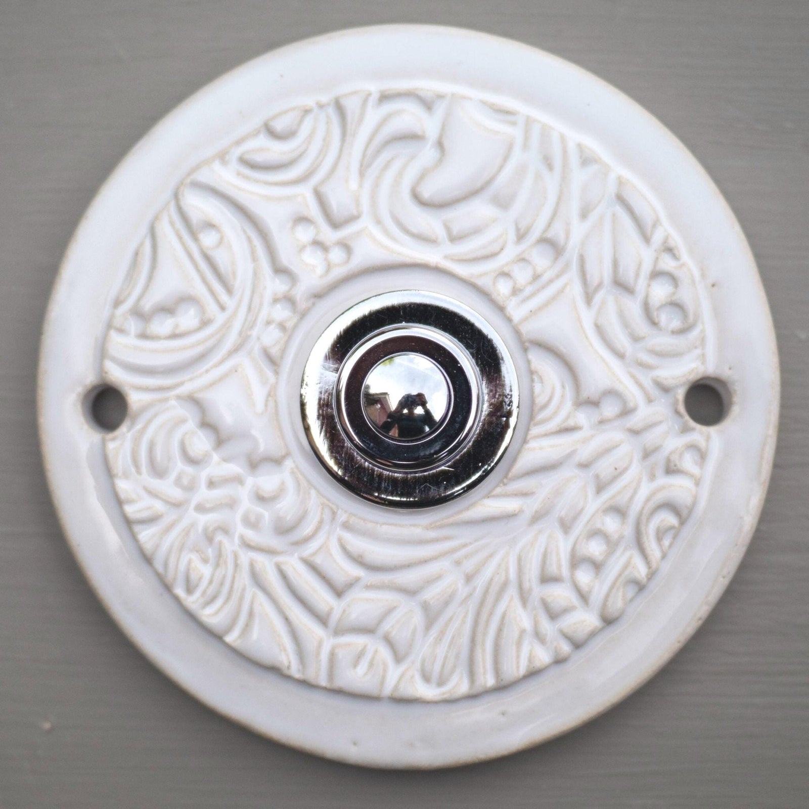 Runde dekorative Klingel mit reinweißem Ornamentmuster auf grauer Platte und goldenem Druckknopf in der Mitte