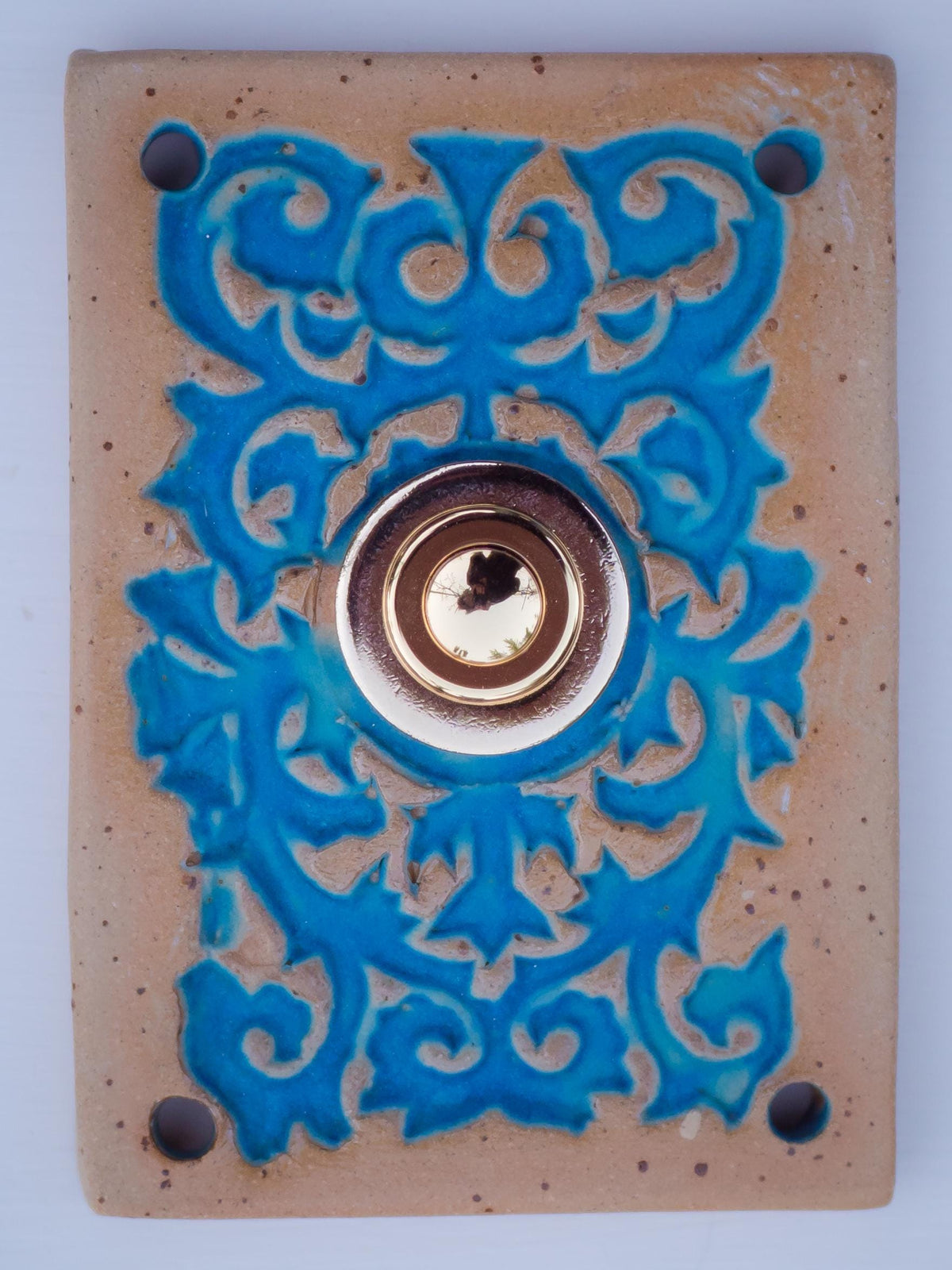 Rechteckige Keramikklingel mit goldbrauner Grundfläche, leuchtend blauem Reliefmuster und zentralem goldenem Druckknopf.