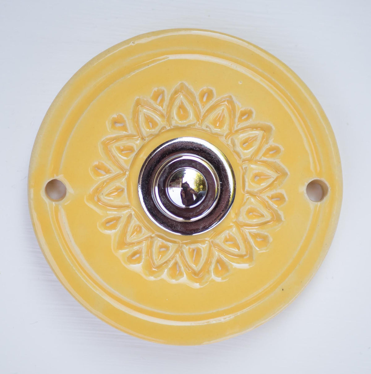 Runde dekorative Klingel mit geprägtem Blumen-Sternmuster auf gelber Platte und silbernem Druckknopf in der Mitte.