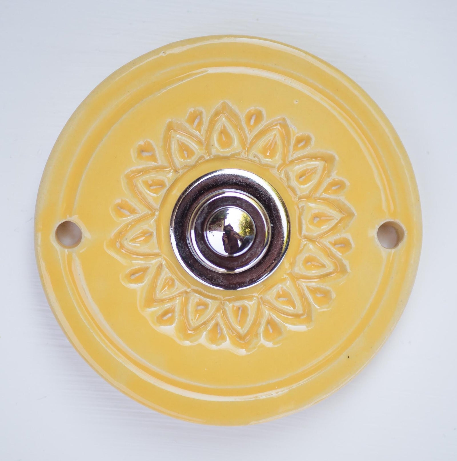 Runde dekorative Klingel mit geprägtem Blumen-Sternmuster auf gelber Platte und silbernem Druckknopf in der Mitte.