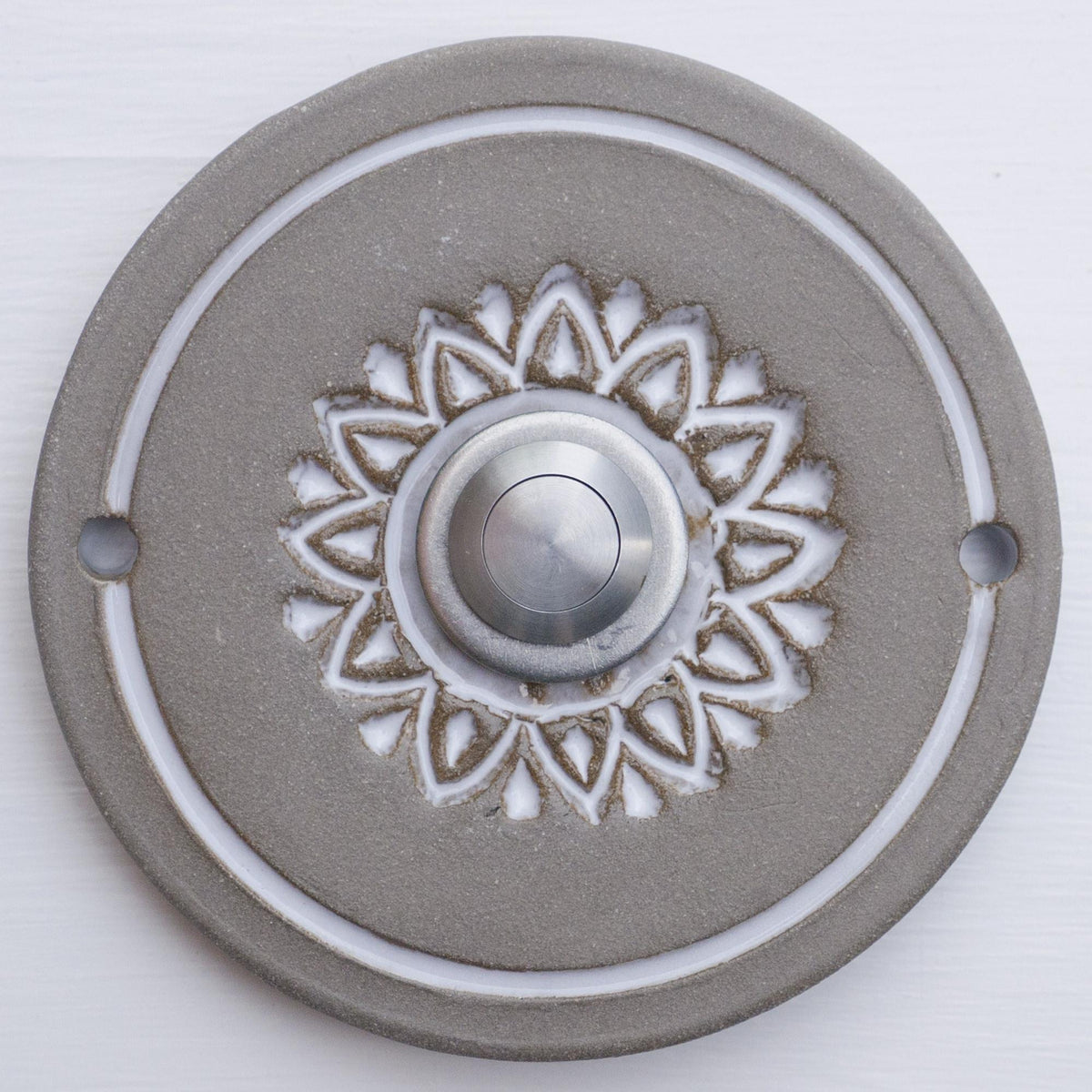 Runde dekorative Klingel mit weißem geprägtem Blumen-Sternmuster auf grauer Platte und silbernem Druckknopf in der Mitte.