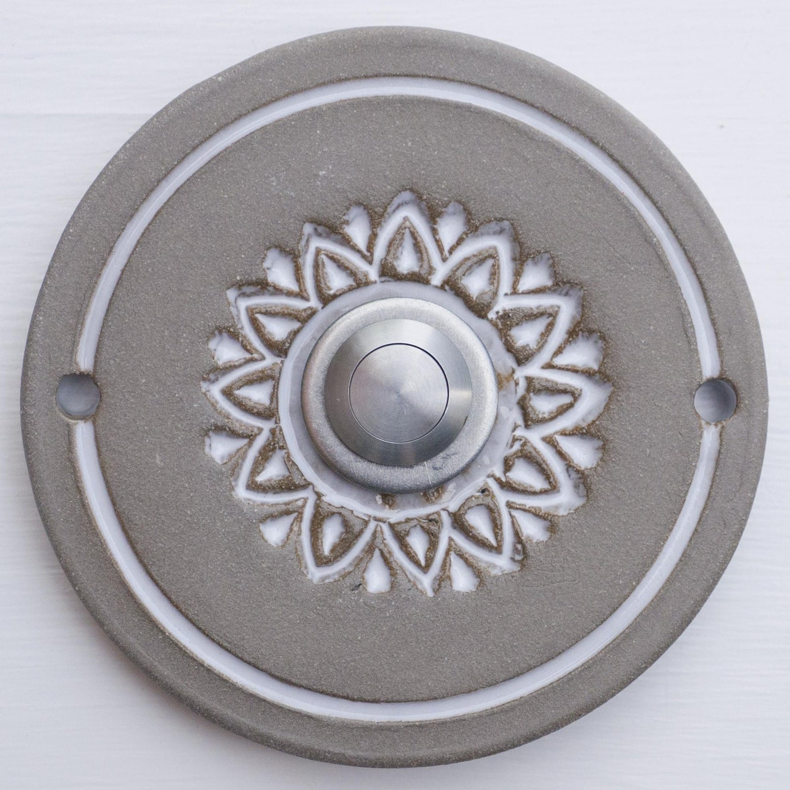 Runde dekorative Klingel mit weißem geprägtem Blumen-Sternmuster auf grauer Platte und silbernem Druckknopf in der Mitte.