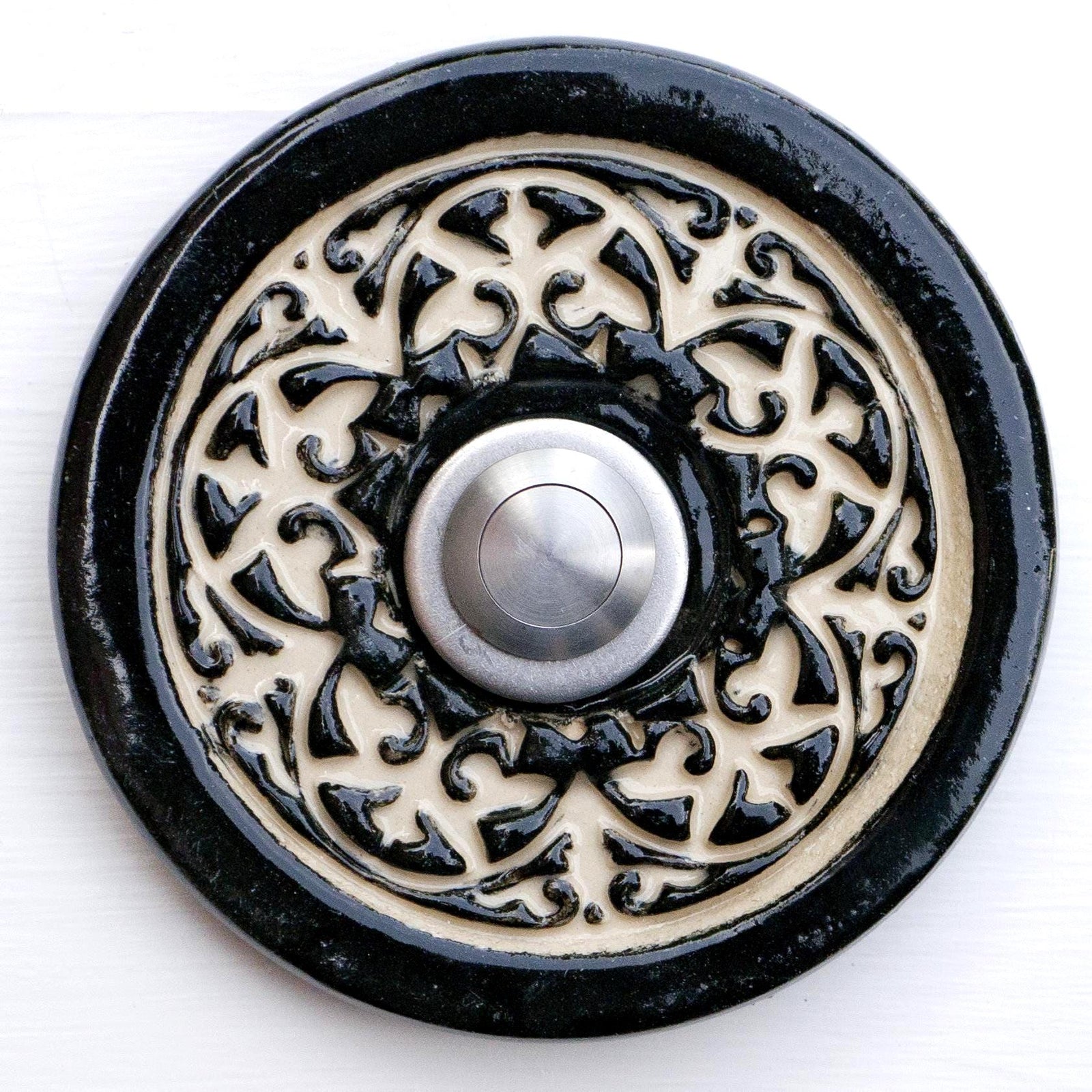 Handgefertigte runde Keramiktürklingel in tiefem Schwarz mit kunstvollem cremefarbenem Reliefmuster. Der Edelstahlknopf setzt einen modernen Akzent in diesem eleganten Handwerksdesign.
