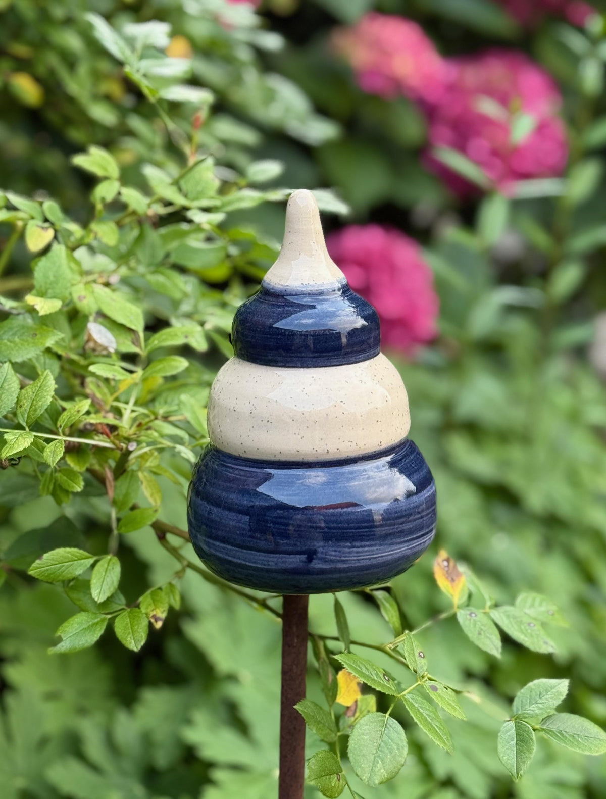 Handgefertigte Keramik-Gartenstele in Blau und Beige, auf einer Metallstange zwischen grünen Pflanzen und pinken Blüten