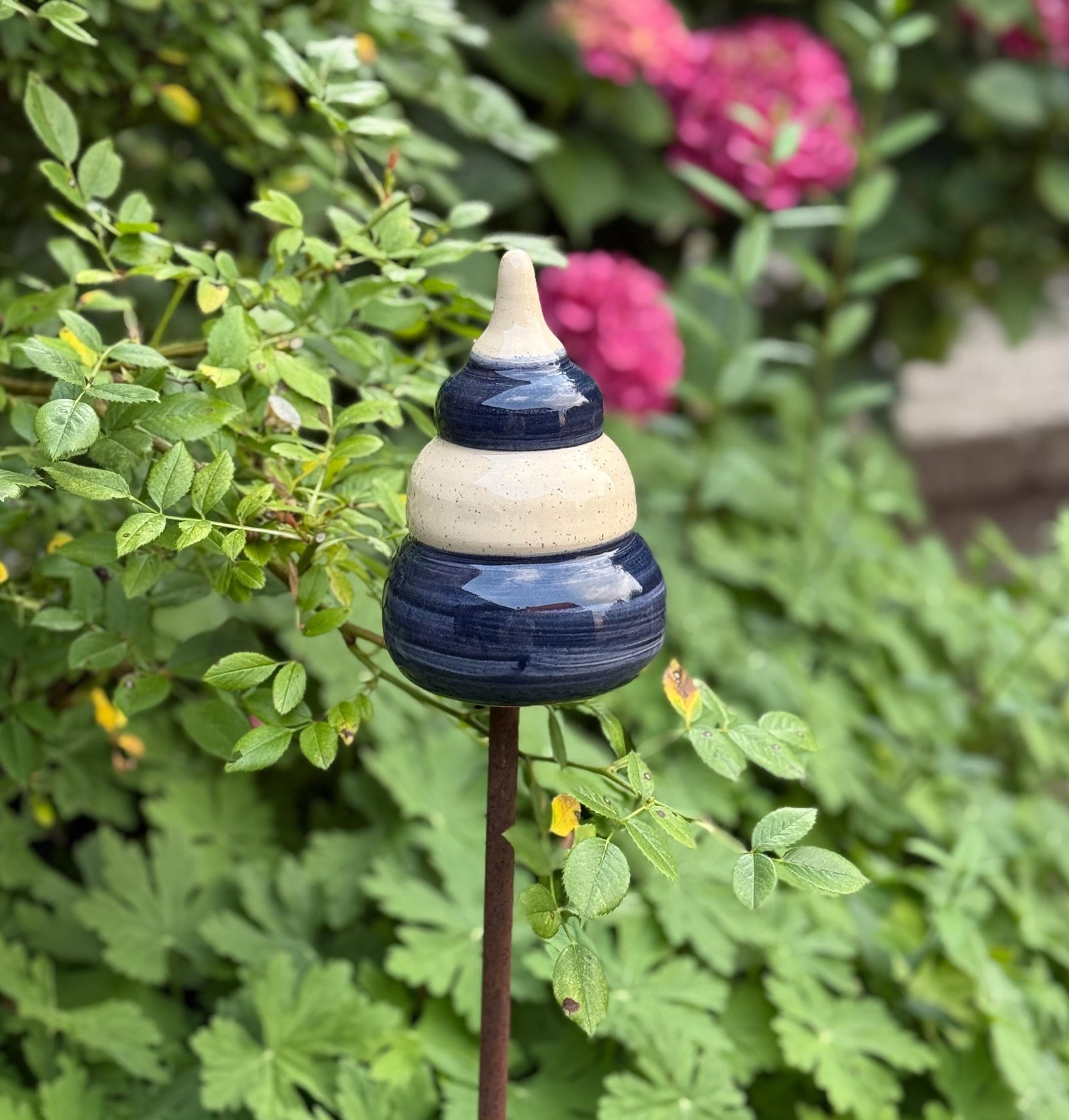 Handgefertigte Keramik-Gartenstele in Blau und Beige, auf einer Metallstange zwischen grünen Pflanzen und pinken Blüten