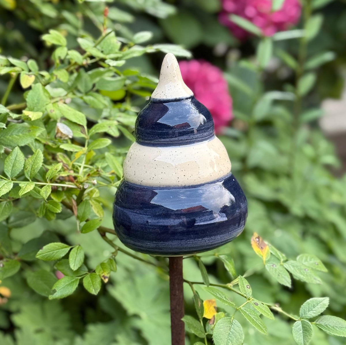 Handgefertigte Keramik-Gartenstele in Blau und Beige, auf einer Metallstange zwischen grünen Pflanzen und pinken Blüten