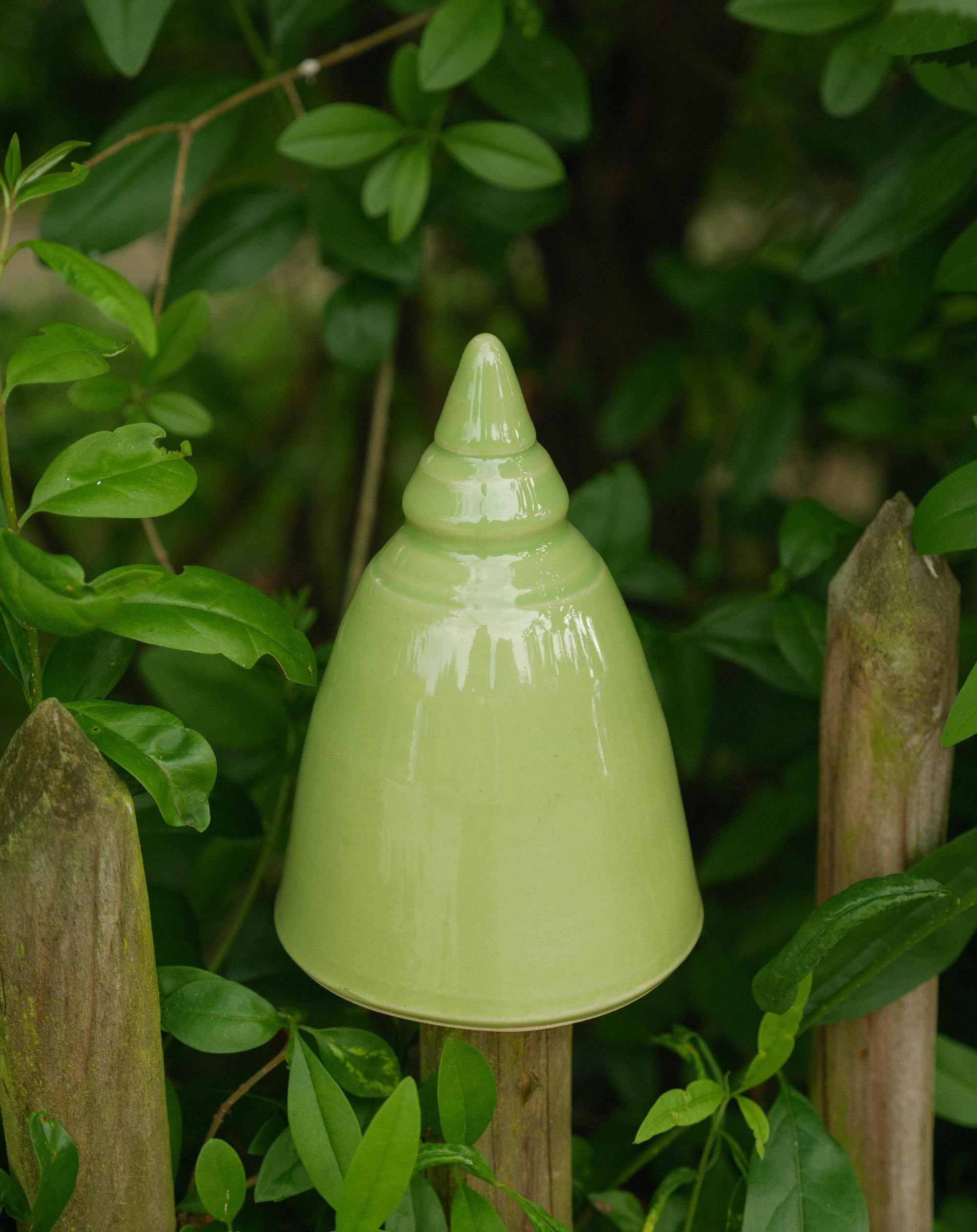 Handgefertigte Keramik-Glocke in glänzendem Hellgrün, auf einem Holzpfosten im Garten befestigt und von grünen Blättern umgeben.