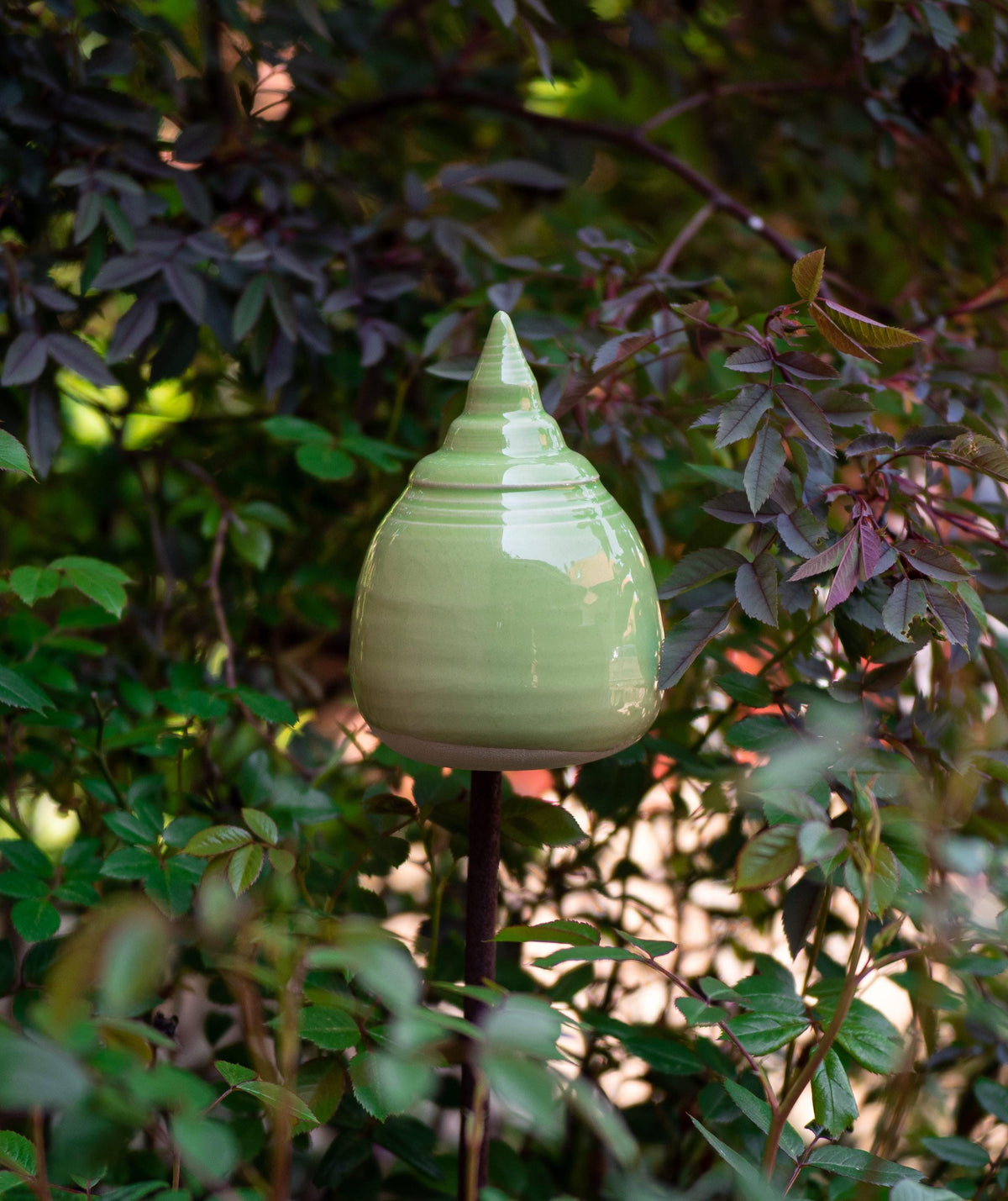 Handgefertigte Keramik-Glocke in abgerundeter Kegelform mit glänzender hellgrüner Glasur, auf einem Metallstab zwischen Blättern befestigt.