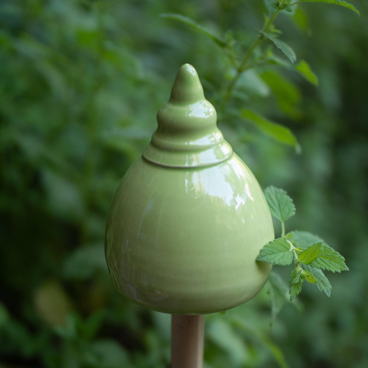 Handgefertigte Keramik-Glocke in abgerundeter Kegelform mit glänzender hellgrüner Glasur, auf einem Metallstab zwischen Blättern befestigt.