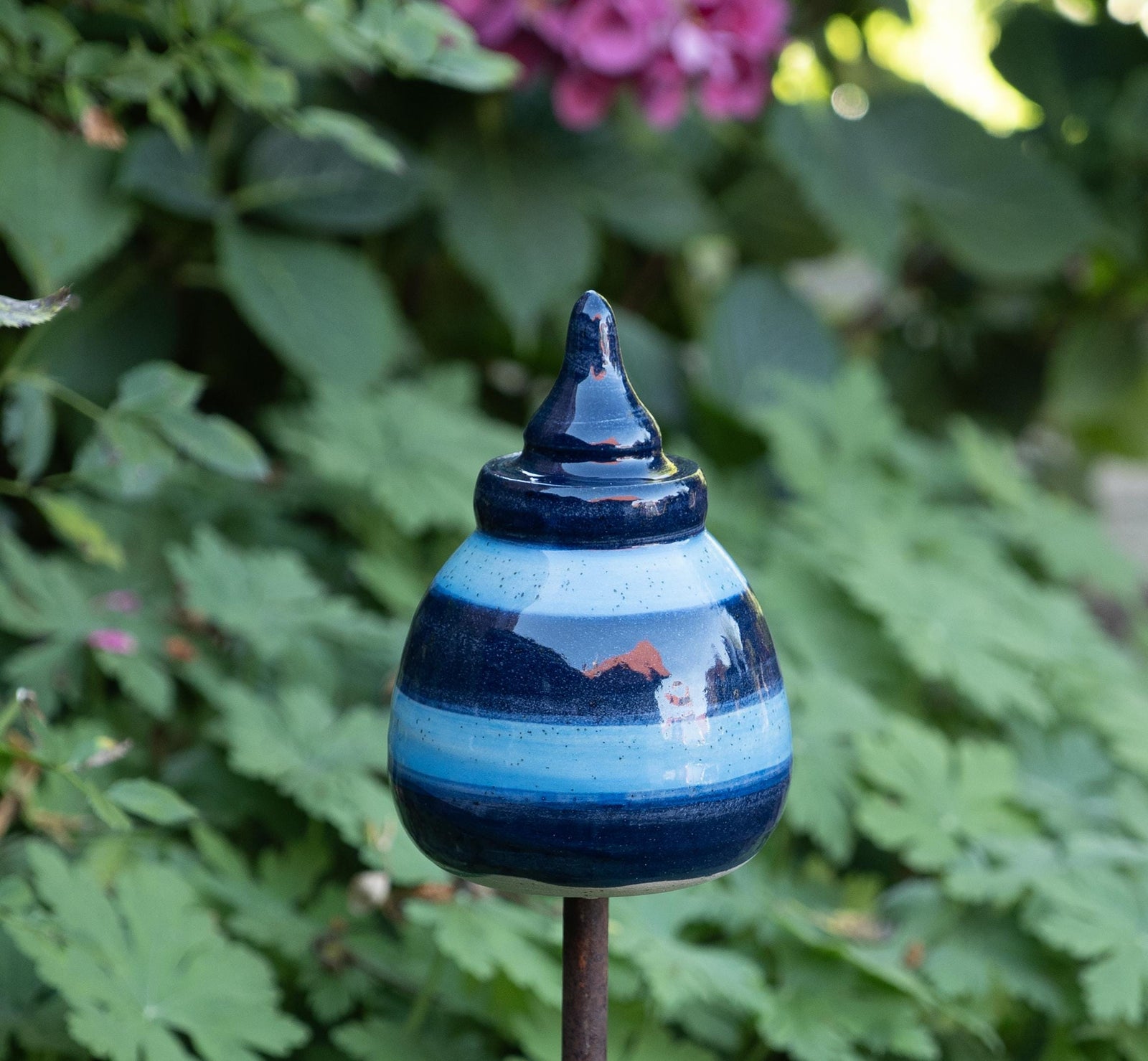 Handgefertigtes Keramik-Gartenornament in abgerundeter Kegelform mit glänzender dunkelblau-hellblauer Streifenglasur, auf einer Metallstange zwischen grünem Laub befestigt