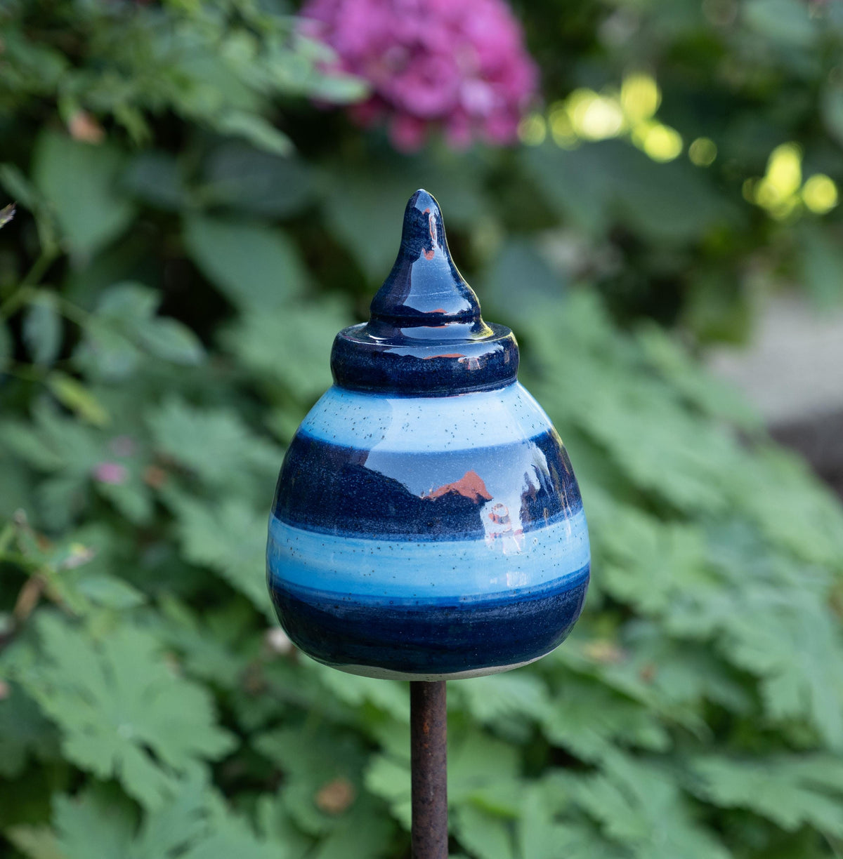 Handgefertigtes Keramik-Gartenornament in abgerundeter Kegelform mit glänzender dunkelblau-hellblauer Streifenglasur, auf einer Metallstange zwischen grünem Laub befestigt