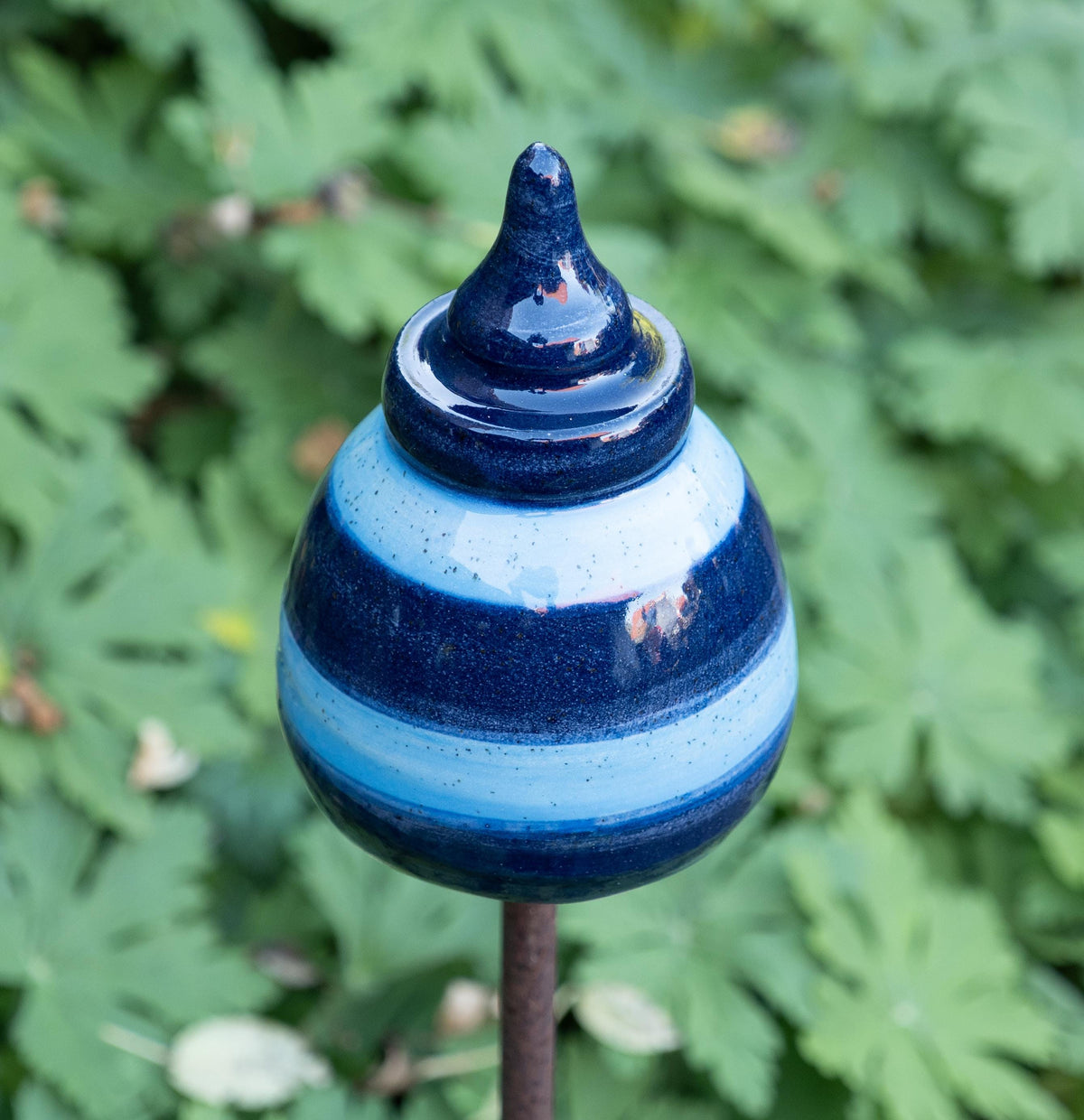 Handgefertigtes Keramik-Gartenornament in abgerundeter Kegelform mit glänzender dunkelblau-hellblauer Streifenglasur, auf einer Metallstange zwischen grünem Laub befestigt