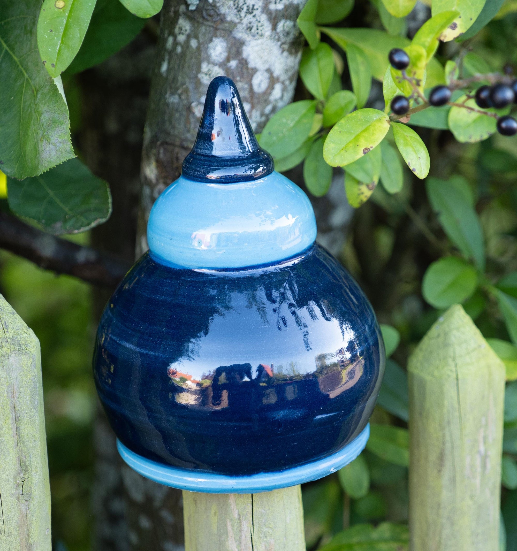Handgefertigte Keramik-Glocke in glänzendem Dunkelblau mit hellblauem Abschluss, auf einem Holzpfosten vor einem Baum mit grünen Blättern und Beeren befestigt.