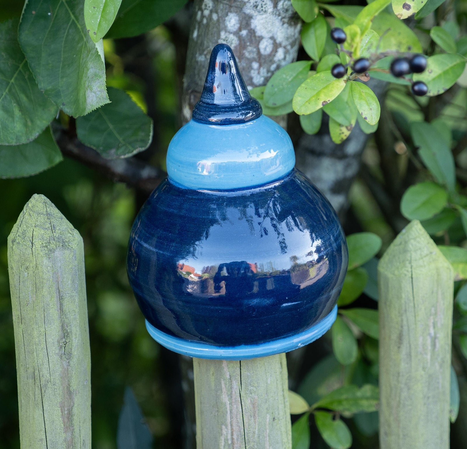 Handgefertigte Keramik-Glocke in glänzendem Dunkelblau mit hellblauem Abschluss, auf einem Holzpfosten vor einem Baum mit grünen Blättern und Beeren befestigt.