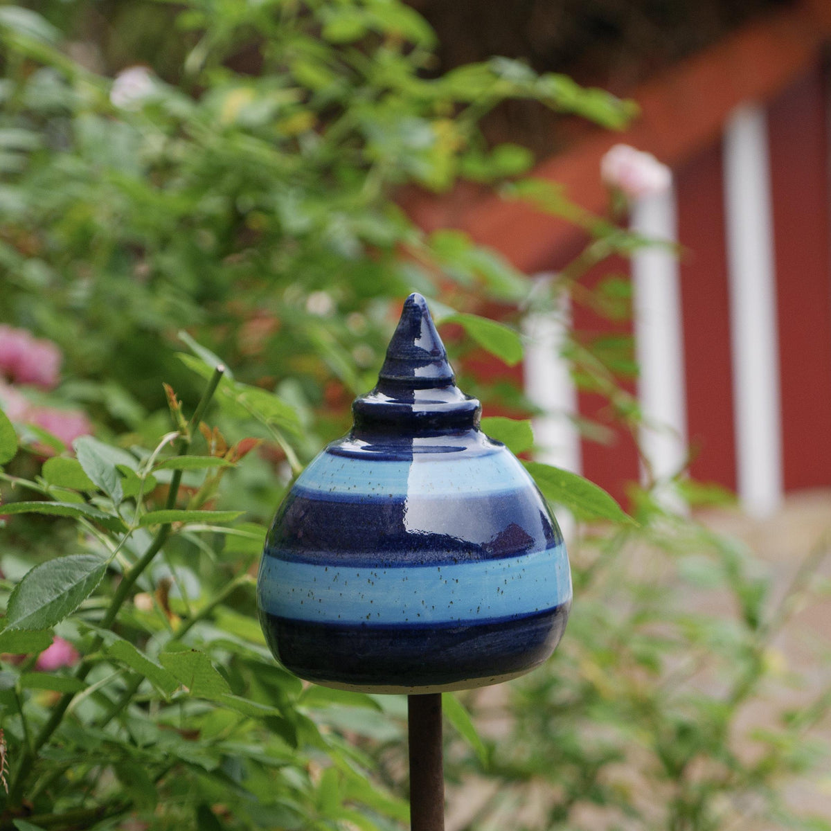 Handgefertigte Keramik-Glocke in konischer Kuppelform mit glänzenden dunkel- und hellblauen Streifen, auf einem Holzstab zwischen grünen Pflanzen befestigt