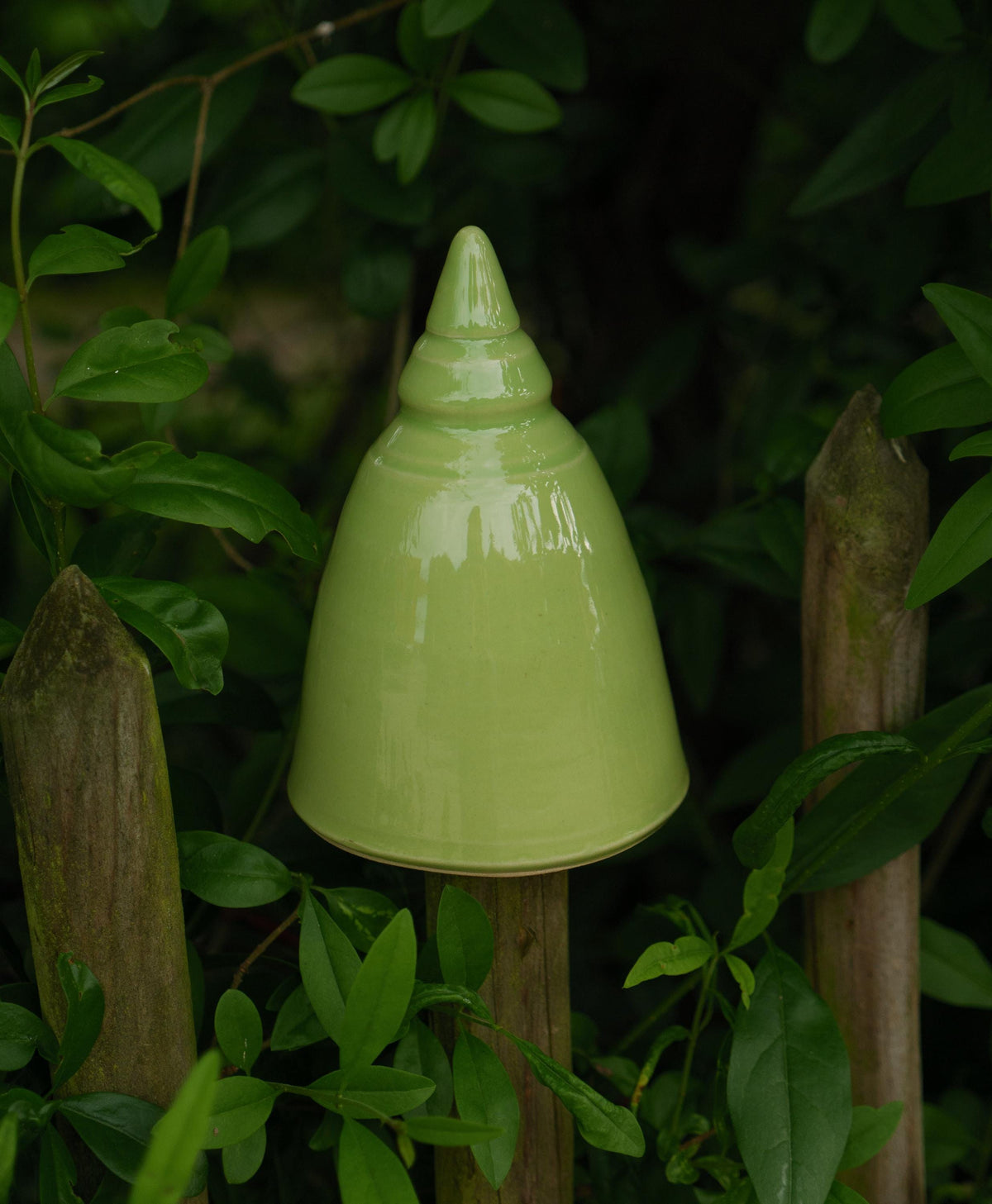 Handgefertigte Keramik-Glocke in glänzendem Hellgrün, auf einem Holzpfosten im Garten befestigt und von grünen Blättern umgeben.