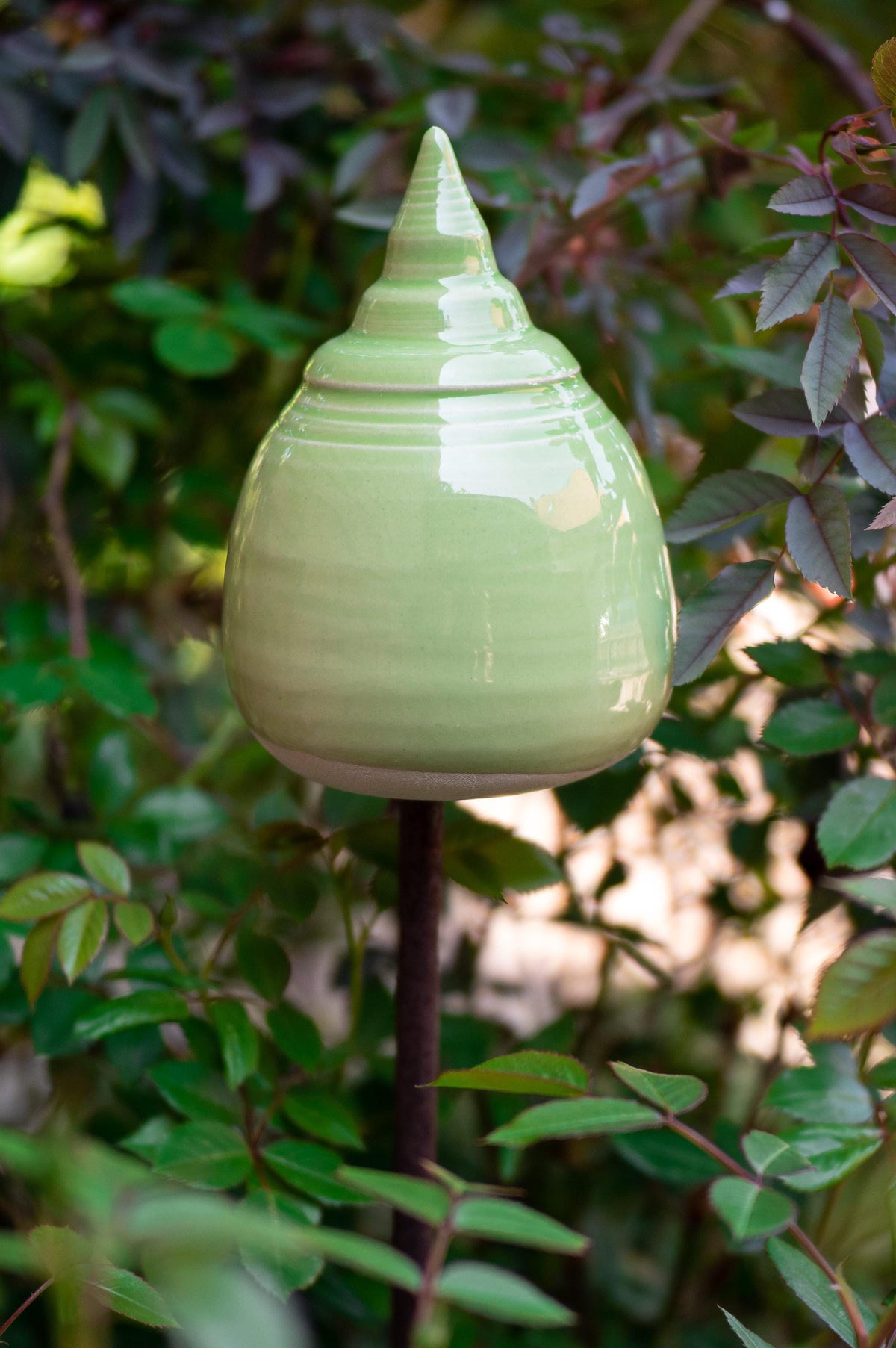 Handgefertigte Keramik-Glocke in abgerundeter Kegelform mit glänzender hellgrüner Glasur, auf einem Metallstab zwischen Blättern befestigt.