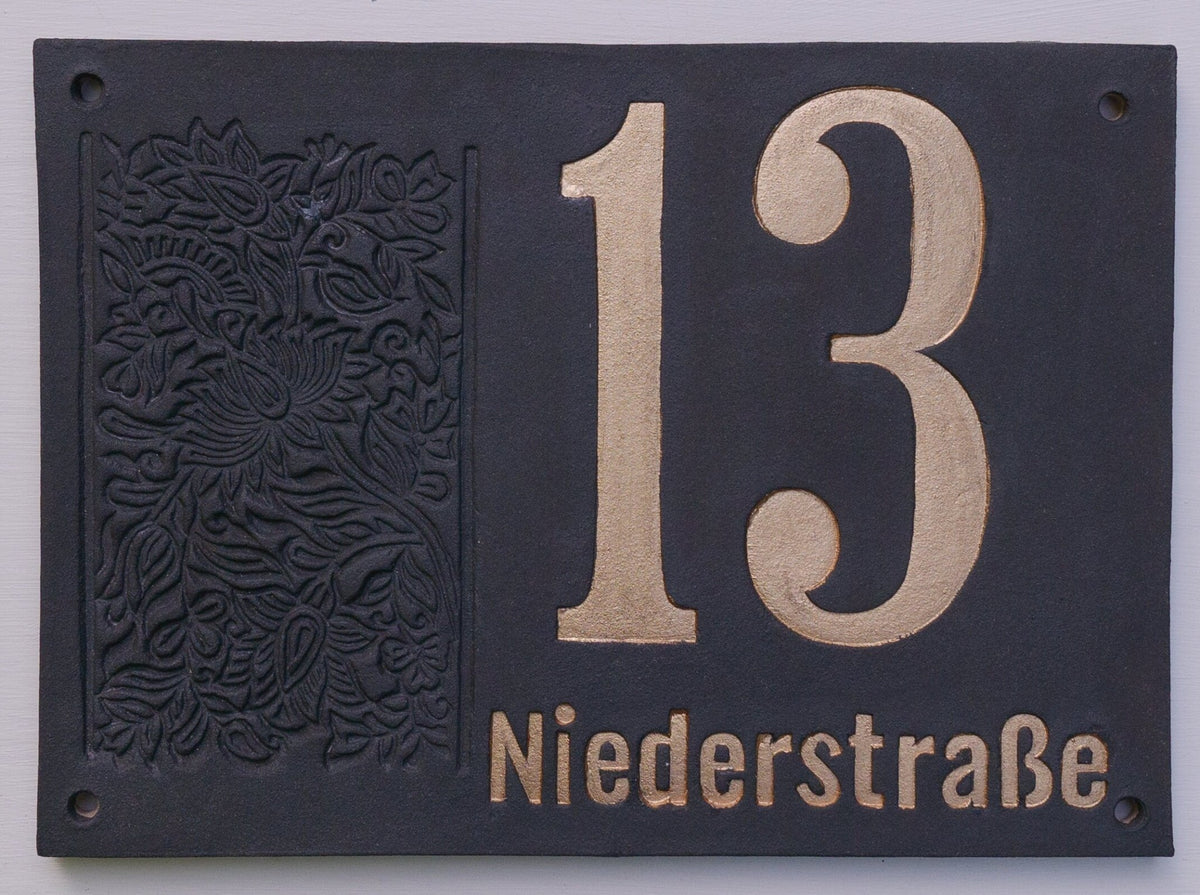 Elegantes Hausnummernschild mit geprägtem Blumenmuster, zeigt die Nummer 13 und den Straßennamen Niederstraße in Gold auf dunklem Hintergrund.