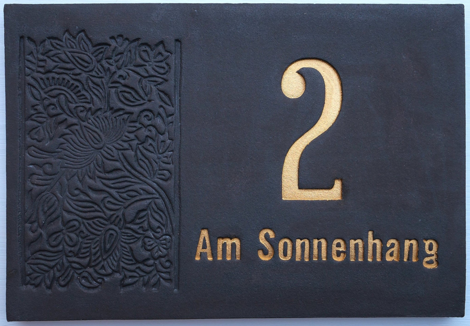 Rechteckiges Keramik-Hausschild in Dunkelblau mit goldfarben eingravierter Zahl 2 und Aufschrift 'Am Sonnenhang', verziert mit floraler Gravur auf der linken Seite
