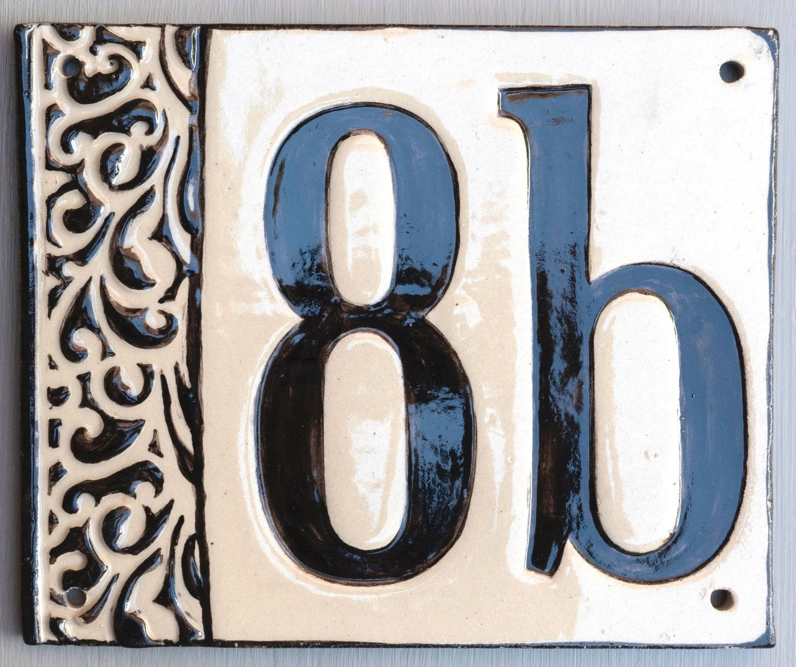 Quadratisches Keramik-Hausschild mit cremefarbenem Hintergrund, glänzenden schwarzen Reliefziffern und Buchstabe 8b sowie erhabenem schwarzem Ziermuster am linken Rand