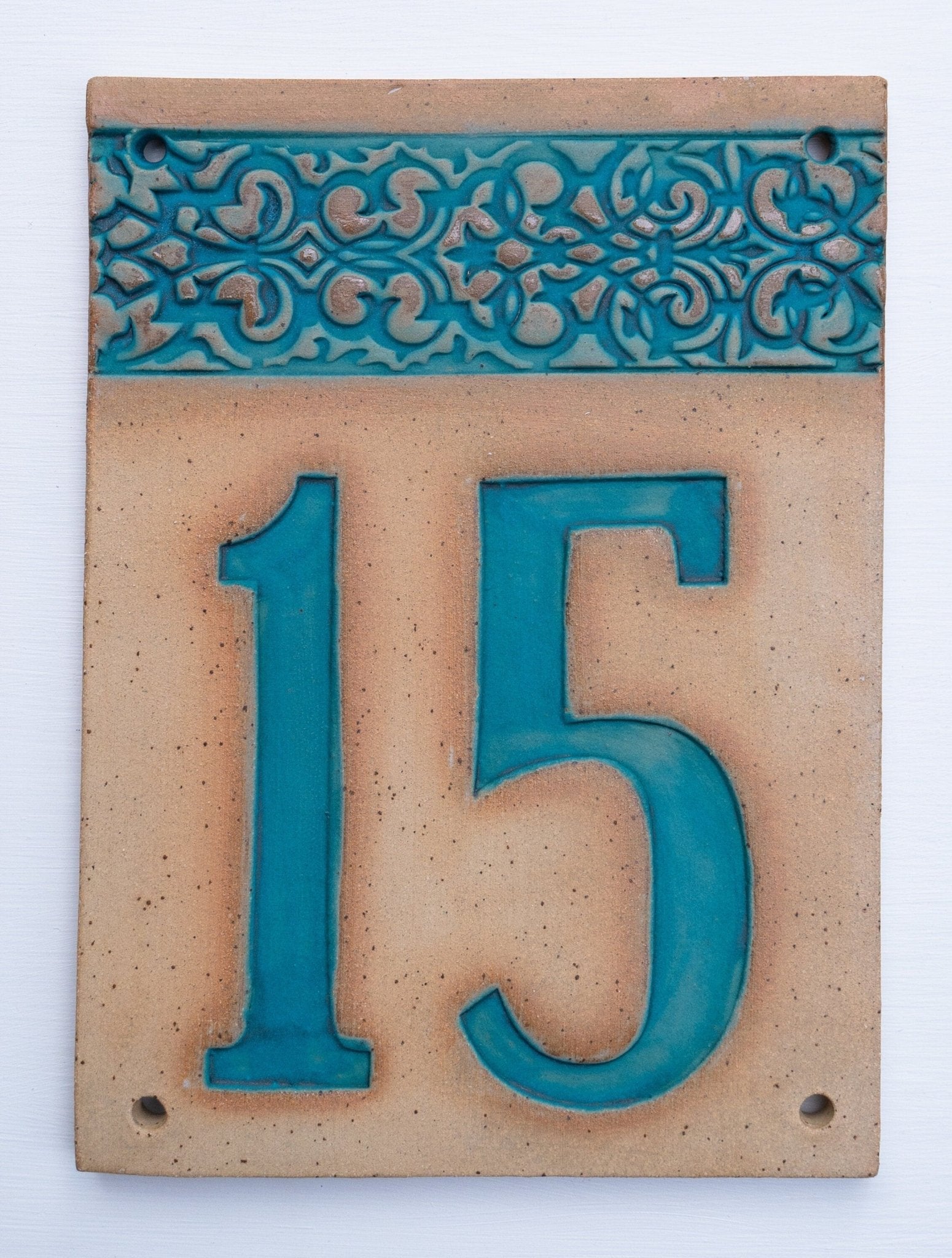 Rechteckiges Keramik-Hausschild mit beigem Hintergrund, türkisfarbenen Reliefziffern 15 und ornamentalem türkisfarbenem Rand auf der linken Seite