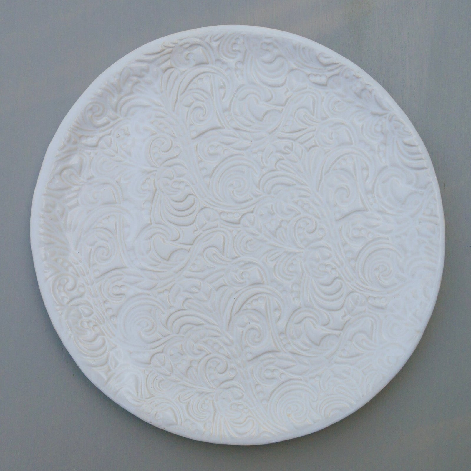 Runder weißer Keramikteller mit geprägtem Blumen- und Wirbelmuster.